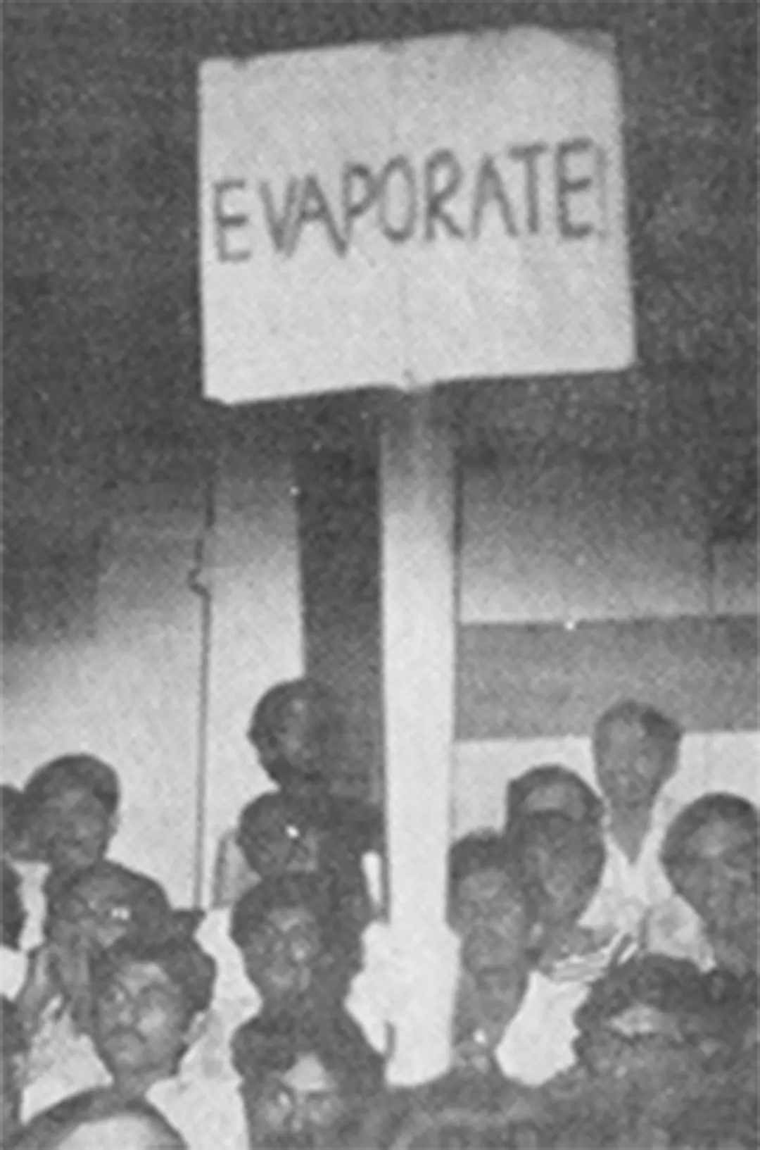 A placard reading EVAPORATE held above a seated crowd