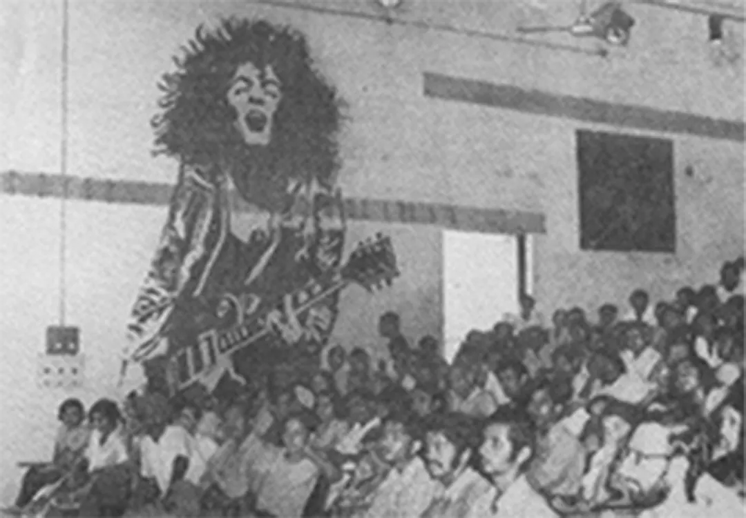 A crowd sits under a giant painted figure on the wall