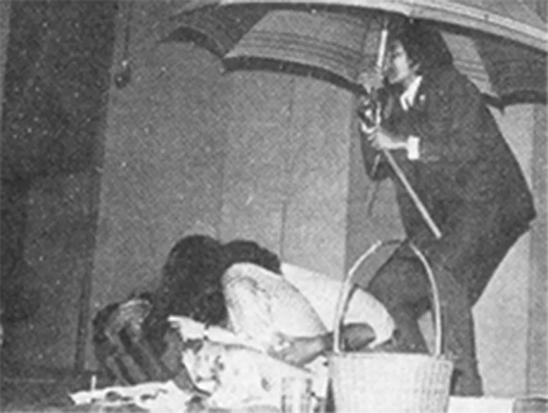 A play: a man in a suit holds an umbrella over a crouching figure