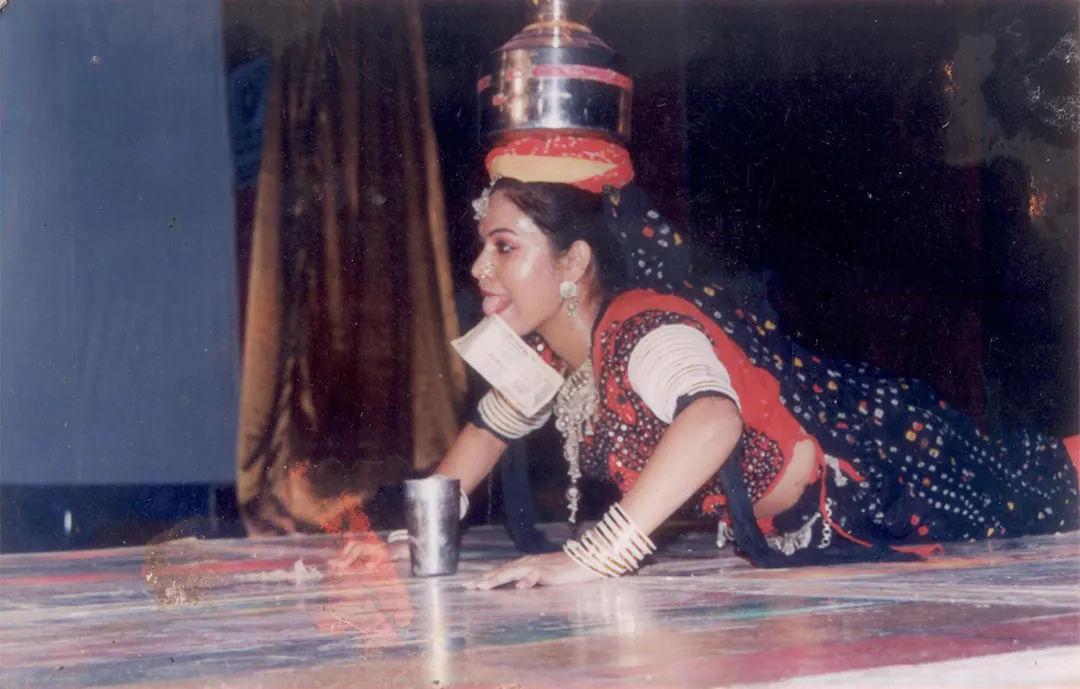 A dancer balances a stack of brass pots on her head, bent low to the stage